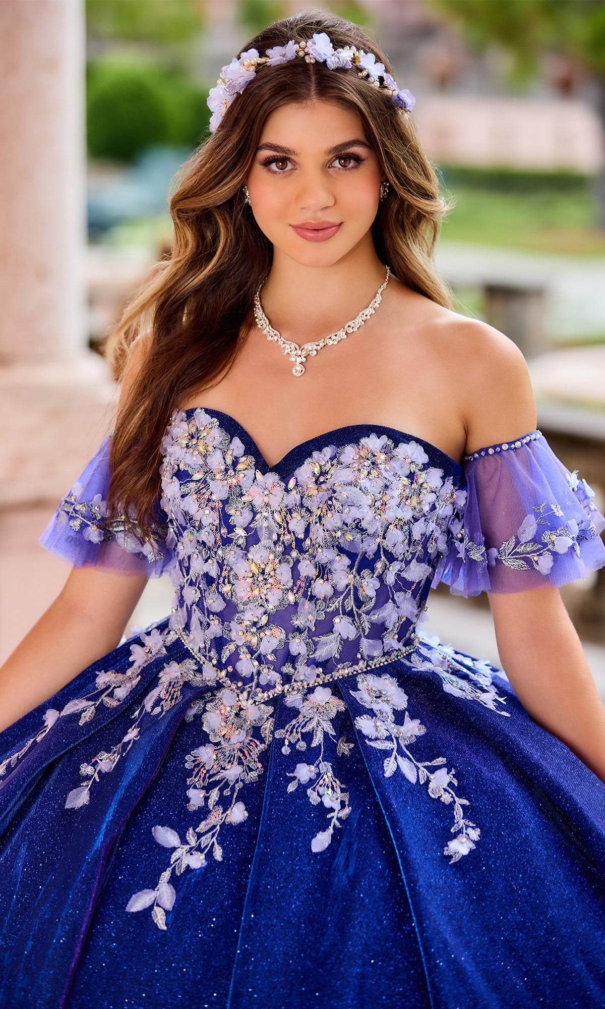 A young woman poses outdoors in the Quinceanera Dress PR30134 by Princesa—a sparkly blue ball gown with floral embroidery, matching headband, and a corset bodice that enhances her enchanting look.
